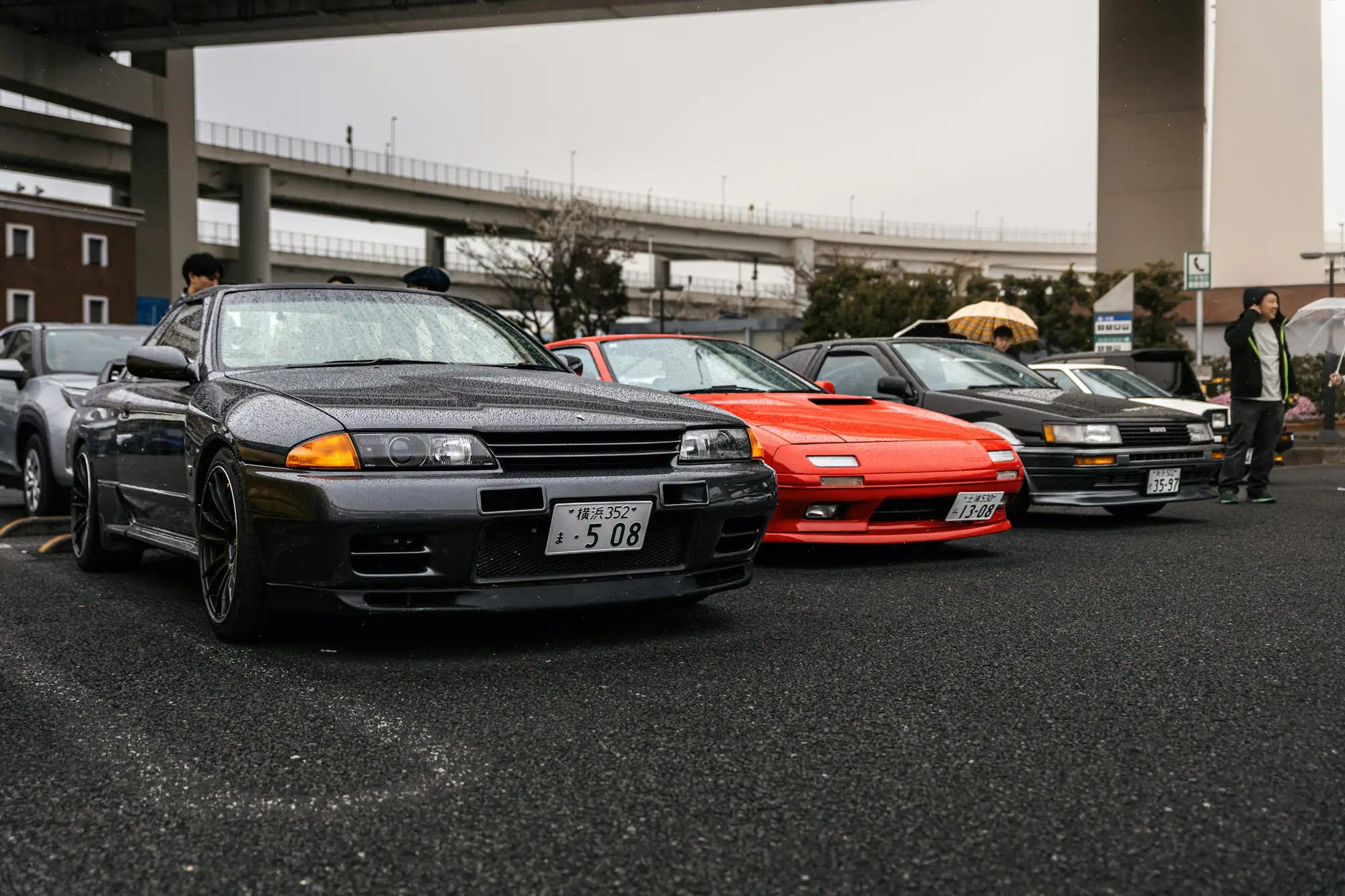 Rows of Japanese-market performance cars parked beneath an expressway in Japan